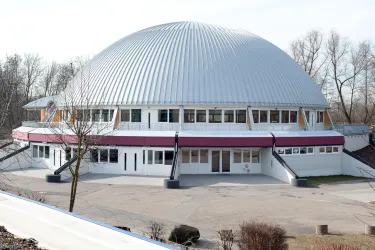 Ein modernes Gebäude mit einer gewölbten Metall-Dachkonstruktion. Es befindet sich in einem offenen Bereich mit Bäumen im Hintergrund.