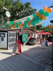 Ein einladender Eingangsbereich mit bunten Luftballons in Orange und Grün. Der Himmel ist klar und es gibt mehrere Stände und Schilder.