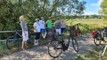Eine Gruppe von Radfahrern steht an einem Weg in der Natur. Im Hintergrund sind Felder und Bäume zu sehen.