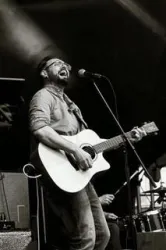 Ein Musiker mit einer Akustikgitarre singt leidenschaftlich auf einer Bühne. Die Aufnahme ist in Schwarz-Weiß und vermittelt eine lebendige Konzertatmosphäre.