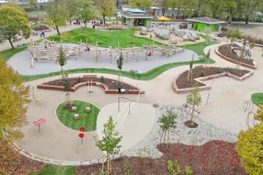 Ein Spielplatz mit verschiedenen Spielgeräten und grünen Flächen. Im Hintergrund sind Bäume und ein Gebäude zu sehen.