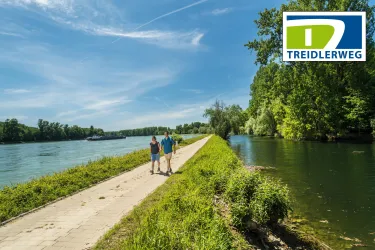 Ein schöner Weg entlang eines Flusses mit grünen Ufern. Zwei Personen spazieren entspannt auf dem Gehweg.