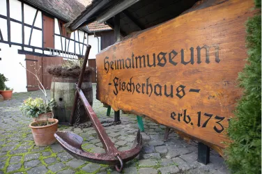 Ein Holzschild mit der Aufschrift "Heimatmuseum - Fischerhaus - erb. 173" steht neben einem Anker. Im Hintergrund sind historische Gebäude sichtbar, die eine ländliche Atmosphäre schaffen.