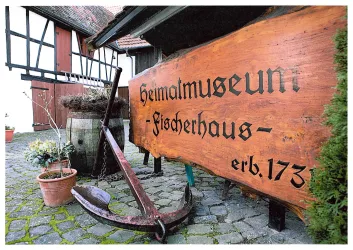 Ein rustikales Schild mit der Aufschrift „Heimatmuseum Fischernhaus“ und einem Anker. Im Hintergrund sind historische Gebäude und Pflanzen zu sehen.