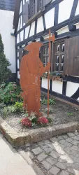 Eine metallene Figur eines Wanderers steht in einem kleinen Garten. Im Hintergrund sieht man ein traditionelles Fachwerkhaus.