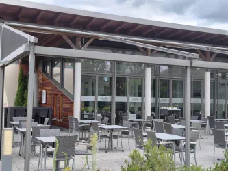 Ein modernes Restaurant mit einer Terrasse und grauen Tischen. Die großen Fenster bieten einen Blick ins Innere.