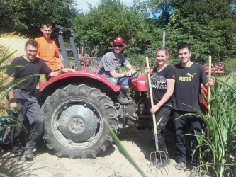 Eine Gruppe von fünf Personen steht lächelnd neben einem roten Traktor. Sie halten Werkzeuge und befinden sich in einer ländlichen Umgebung mit viel Grün.