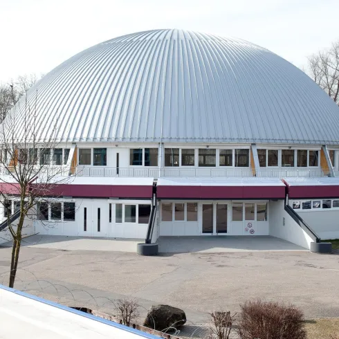 Ein modernes Gebäude mit einer gewölbten Metall-Dachkonstruktion. Es befindet sich in einem offenen Bereich mit Bäumen im Hintergrund.