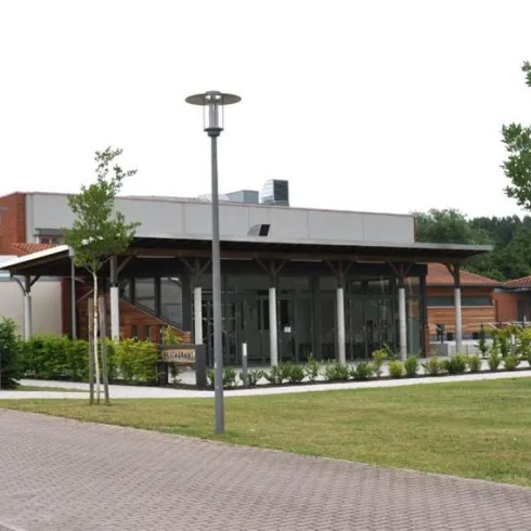 Ein modernes Gebäude mit einer überdachten Terrasse und vielen Bäumen im Vordergrund. Die Umgebung ist gepflegt und lädt zum Verweilen ein.